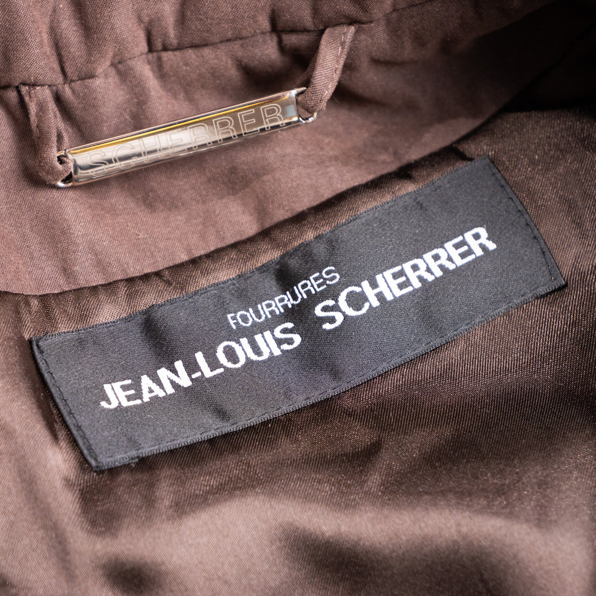 Close-up interior view of the woven black Fourrures Jean-Louis Scherrer main brand label and silver metal nameplate hanging chain.
