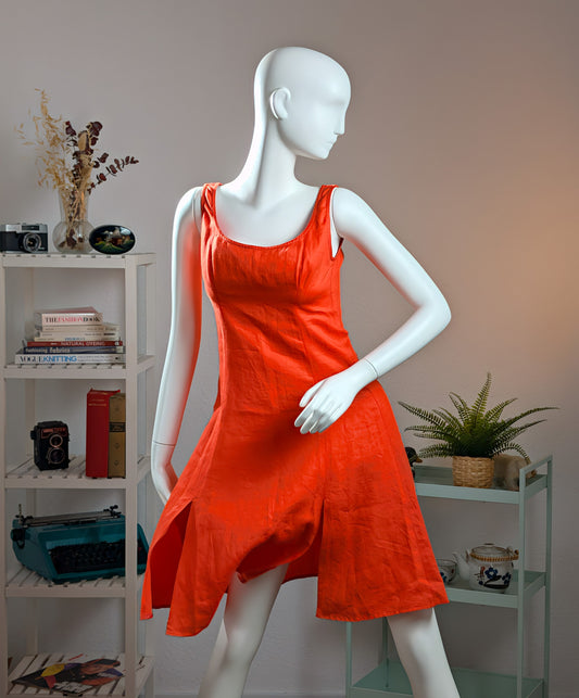 Front view of an authentic Gianni Versace Spring/Summer 1993 runway prototype dress worn by Emma Sjöberg displayed on a white mannequin. The knee-length, sleeveless dress is crafted from vibrant orange-red linen. It features a form-fitting bodice with a scoop neckline, structured princess seams, and two distinct front slits on the flared A-line skirt. The background shows a minimalist studio setting with a white bookshelf.