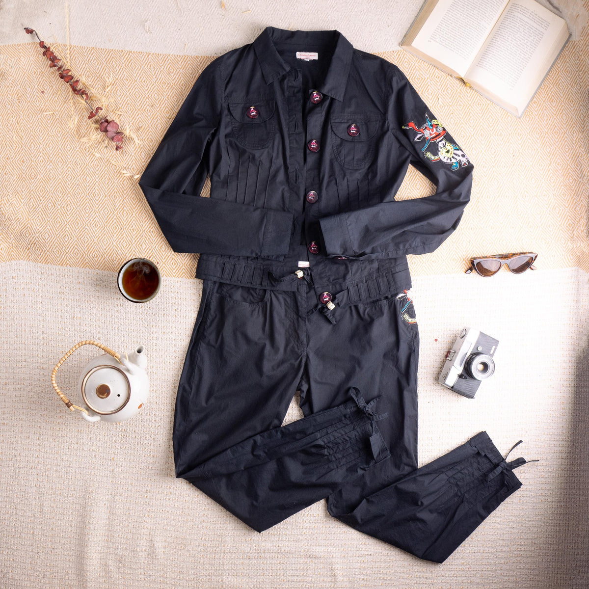 Pristine Deadstock Christian Lacroix Archival Early 2000s Embroidered Cotton Utility Set – jet black crisp cotton, cropped utility jacket with point collar, five dark red enamel logo buttons, two chest flap pockets, fringe-cut hem detail, multicolour floral embroidery patch on left sleeve; matching high-waist cropped tapered trousers with pleated waistband, drawstring tie, fringe-cut hem and matching embroidery at hip, flat lay editorial shot