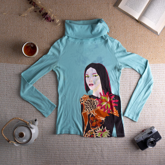 Christian Lacroix Bazar 90s Archival Pop Art Face Print Turtleneck Top – aqua blue stretch jersey, oversized cowl turtleneck, long sleeves, large-scale pop art illustration of a dark-haired woman with bold green eye makeup holding tropical flowers and leopard print fabric, rhinestone embellishment detail, art-to-wear collector piece, flat lay editorial shot